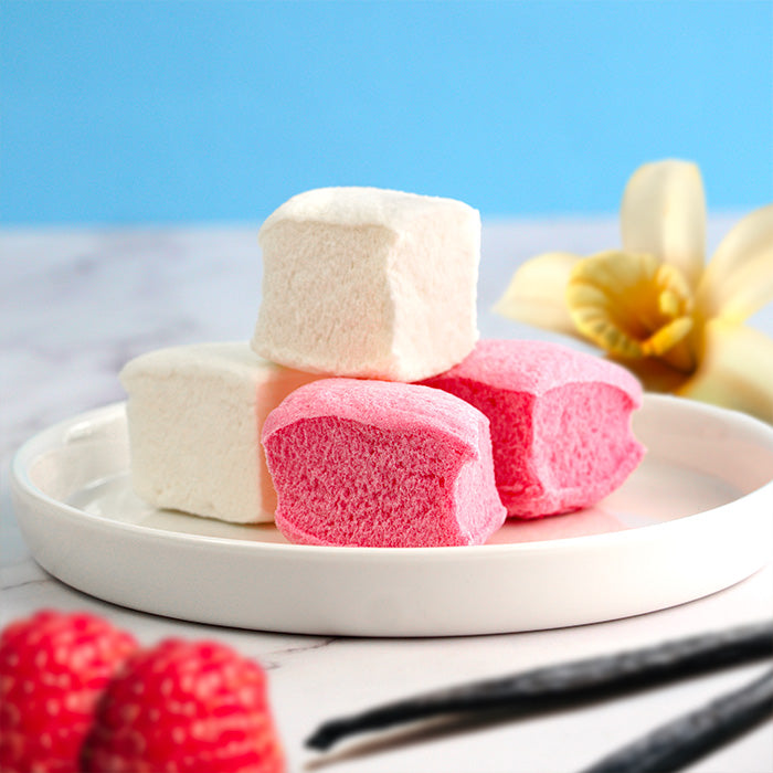 Guimauves blanches et roses dans une coupelle plate sur marbre, avec fleur, gousse de vanille et framboise floutées en arrière-plan