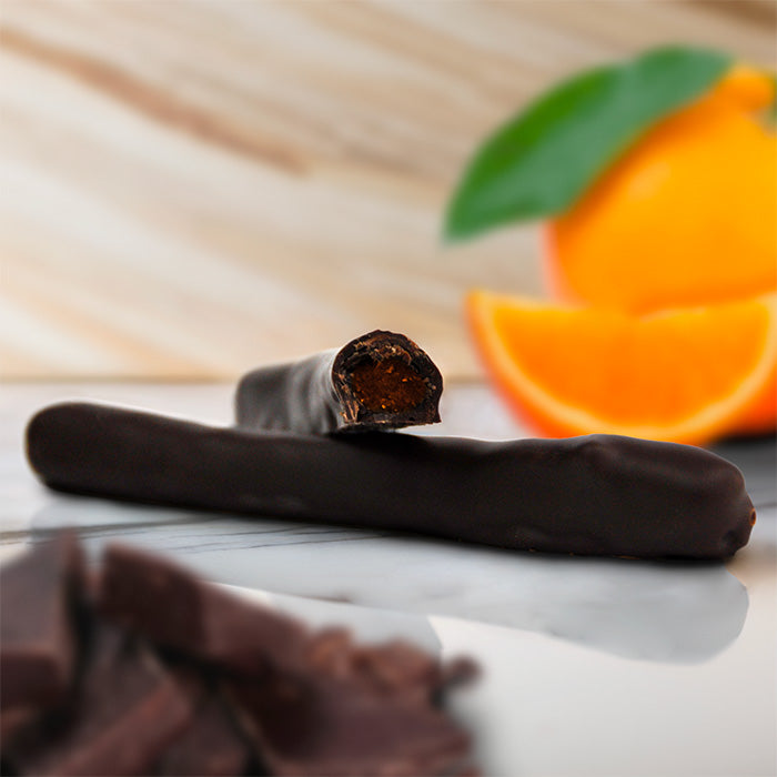 Orangette artisanale en gros plan, une pièce coupée révélant l'intérieur, orange entière en fond et copeaux de chocolat floutés sur marbre