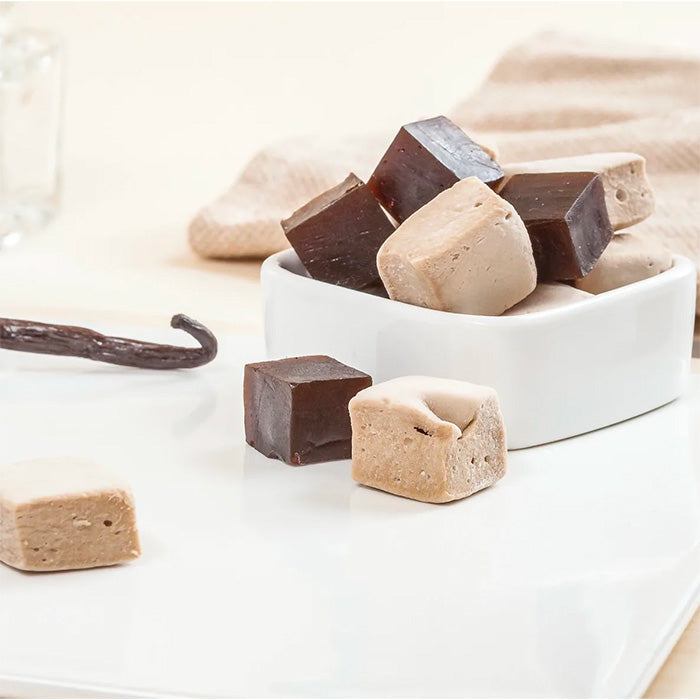 Duo de Réglisse : cubes de réglisse et réglisse à la vanille dans une coupelle avec gousse de vanille, sur table blanche et serviette beige